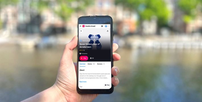A person holds a smartphone displaying a tour booking page for a 'Queer history tour of Amsterdam' from the website 'timetotravel', with a blurred canal and trees in the background.
