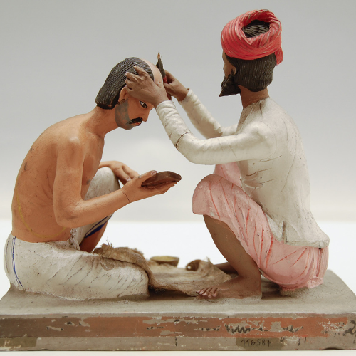 A painted figurine depicts a traditional Indian barber shaving a seated mans head. The barber wears a red turban and white clothing; the client holds a small mirror and wears a white cloth around his waist.