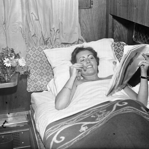 A woman lies in bed inside a train compartment, smiling as she eats grapes and reads a magazine. A blanket covers her, and there are flowers on the table beside her.