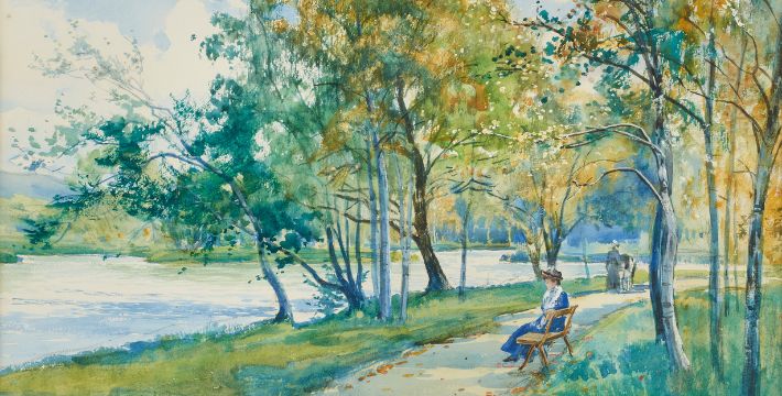 A woman sits on a bench near a river under leafy trees, while two people walk along the path in the background on a sunny day.