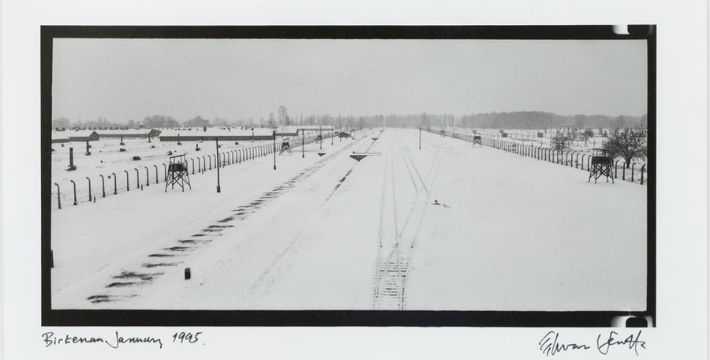 Black and white photo of snow-covered railway tracks leading into Auschwitz-Birkenau concentration camp, bordered by barbed wire fences and guard towers. The landscape is flat and desolate under a winter sky.