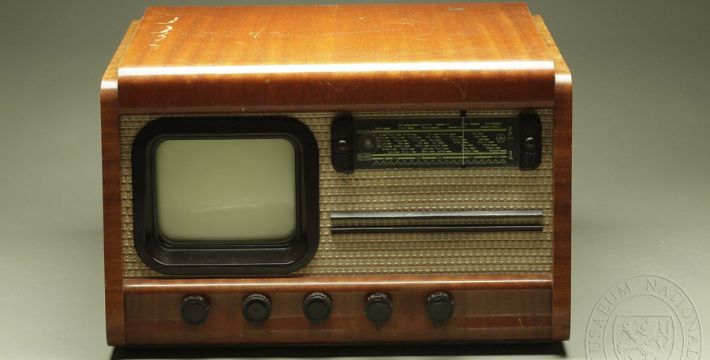 A vintage wooden television set with a small screen on the left, a speaker grille in the center, dials and knobs along the bottom, and a row of tuning indicators on the right.