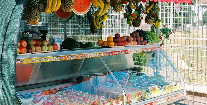 A colorful outdoor fruit stand displaying fresh whole fruits and neatly cut fruit cups inside a refrigerated glass case. Bananas, pineapples, and watermelons hang decoratively from the canopy above. A metal fence is visible in the background.