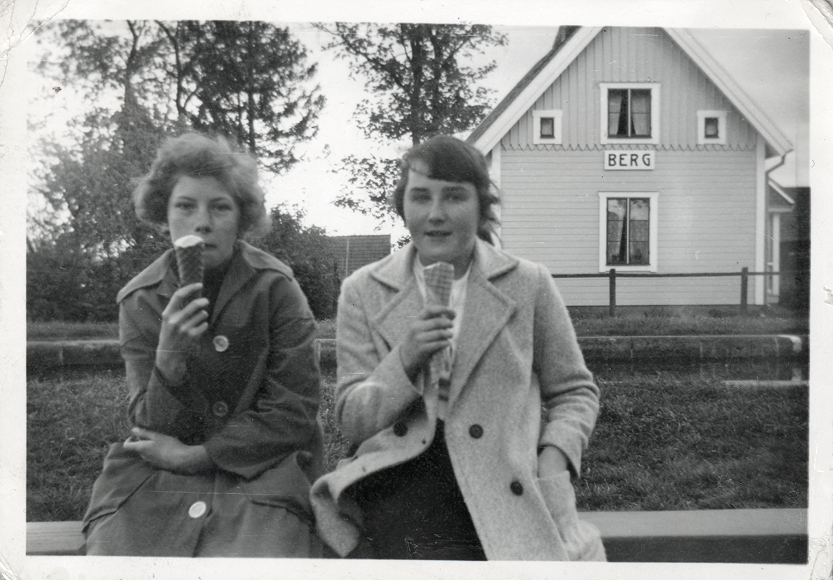 Black and white photo of two women eating ice cream from a cone, looking at the camera. They're wearing coats. There are some trees in the background and a wooden hut carrying the sign 'berg'.