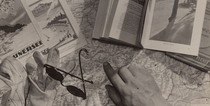 Black and white image depicting travel guides, a pair of gloves and sunglasses, and a hand pointing on a map