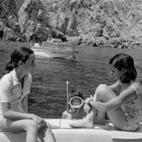 Two women in swimsuits sit on a boat in a rocky coastal area, one adjusting a foot strap. A person in snorkeling gear peeks between them, with a boat in the background.