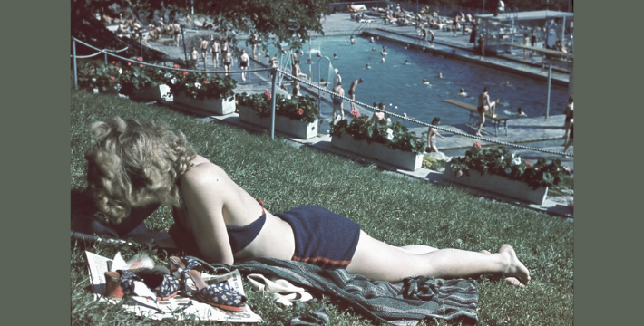 A woman in a blue bikini sunbathes on a grassy hill by a busy pool, with people swimming and lounging around. The scene is relaxed and summery.