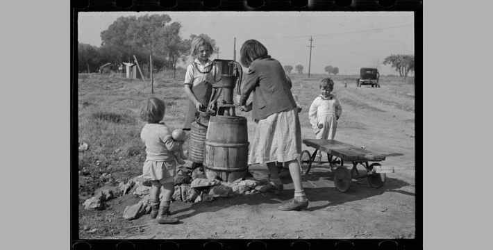 Four children by a water pump, two older children pumping water and the other two younger ones looking on.