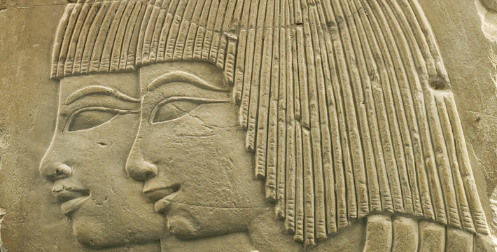 Details from a relief: a gray stone slab on a black background with a relief sculpture of two female faces and necks pointing looking to the left, in Ancient Egyptian style