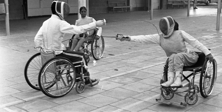 black and white photograph, two wheelchair user athletes in fencing costumes are fencing.