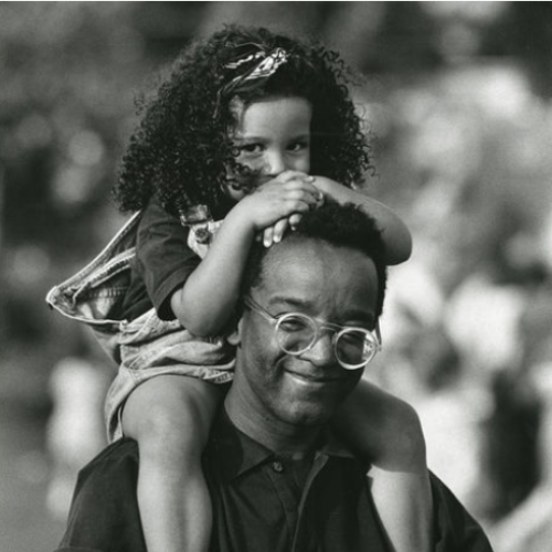 A black man with glasses gives a piggyback ride to a smiling young black girl outdoors.