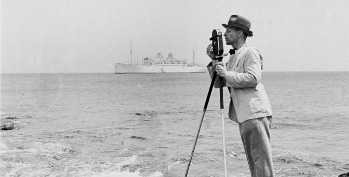A black and white photo of a person stood by the sea with camera on a tripod
