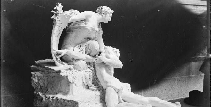 Black and white photo of a sculpture of a woman leaning on one knee holding a man lying on the ground
