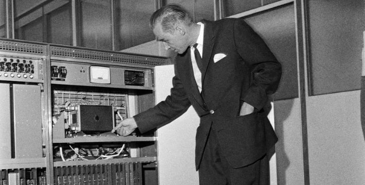 A black and white photo of a man in a suit next to a large computer