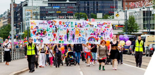 A photo depicting a pride parade with a crowd of people marching with colourful banners and ballooons